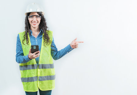 Female builder worker holding phone pointing at blank space isolated. Smiling female engineer holding phone and pointing to the sideの写真素材