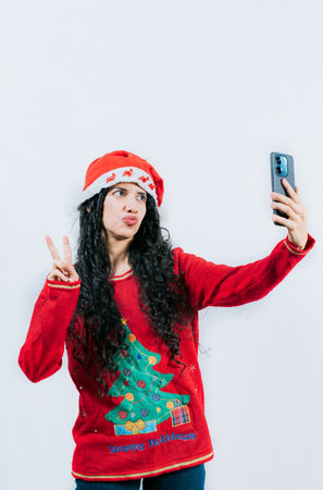 Latin girl in Christmas hat taking selfies and making gestures isolated. Cheerful young woman in Christmas sweater taking selfiesの写真素材