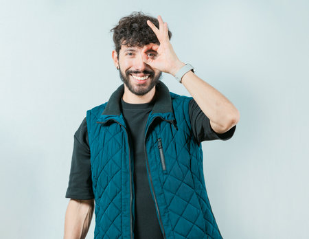 Handsome young man making OK gesture looking through fingers isolated. Cheerful guy making ok...