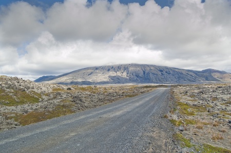 View of southern face of Snaefellsjokull volcano (Iceland). Top of volcano is covered by clouds. A track goes through a lava field covered by mosses and lichens.の写真素材