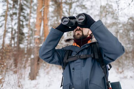 Bearded man using binoculars to orientate while his winter forest trip