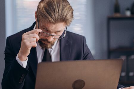 Confused mature business man in black suit and eyeglasses sitting at table and looking at computer screen. Professional office manager using laptop at workplace.の写真素材