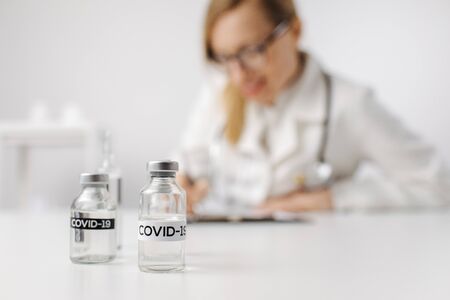 Close up of test tubes with name of COVID-19 standing on table with blur background of female doctor that writing database in clipboard. Concept of vaccine and health treatment.の写真素材