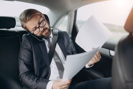 Bearded businessman in formal clothing having online consultation with private lawyer while riding car. Mature man in eyeglasses talking on smartphone and looking through new business contract.の写真素材