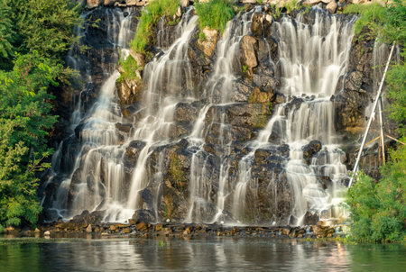 Artificial waterfall above river in summer day. Water cascade flowing on stones. Splash stream between the trees. Cold current descends on the shore. City recreation park.の写真素材