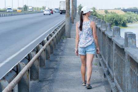 Woman walks road bridge and thoughtfully looks at picturesque river. Walking on the pedestrian zone of the bridge between the highway and iron-concrete handrail.の写真素材