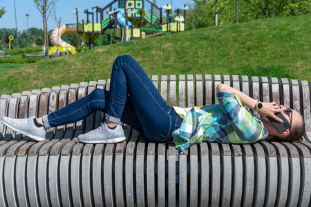 Woman lying down on park bench and talking on mobile phone. Young adult use smart phone for chatting. Female resting and conversation on cell phone. Leisure, technology and modern.の写真素材