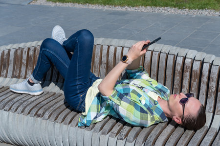 Woman lying down on park bench and flip through display on mobile phone. Young adult use smart phone...
