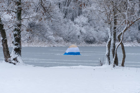 Lonely colormix tent for winter fishing on snow covered in ice river. Fisherman camp nestled in snowy landscape on frozen lake. Ice fishing shelters on cool water in winter.の写真素材