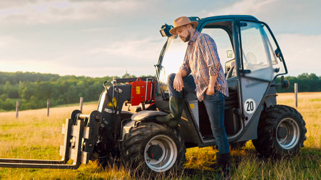 Close up of Caucasian handsome young male farmer in hat standing at tractor, smiling and looking at...