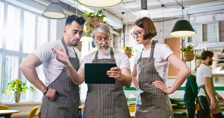 Portrait of Caucasian senior bearded man owner of cafeteria typing on tablet device browsing using app and speaking with young male and female workers. Family business. Restaurant serviceの写真素材