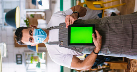 Vertical orientation of happy handsome young Caucasian male employee in medical mask and apron standing in cafe at workplace, looking at camera, holding tablet with green screen Small business conceptの写真素材