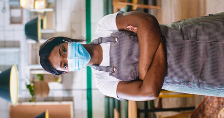 Vertical foto of young beautiful African American woman worker in medical mask and apron standing in coffee house looking at camera indoor. Work in quarantine. Small business concept, portraitの写真素材