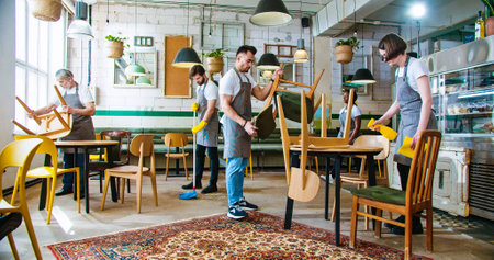 Diverse people workers in coffee house working together putting chairs on tables to clean area at...