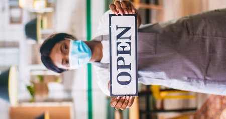 Portrait of pretty young African American young female employee in apron wearing medical mask standing in coffee shop holding in hands Open sign, reopening in quarantine, businessの写真素材