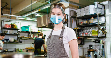 Portrait of cheerful Caucasian young pretty female cafe employee in medical mask wearing face shield at workplace looking at camera standing in restaurant kitchen. Work in virus quarantineの写真素材