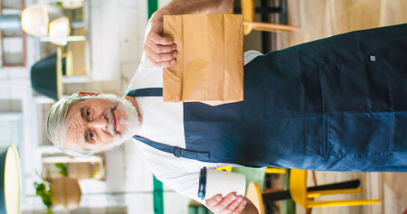 Vertical footage of handsome happy senior positive Caucasian bearded man waiter in apron holding paper bag with take away order and coffee-to-go, looking at camera and smiling, business conceptの写真素材