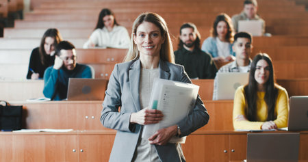 Portrait of beautiful Caucasian female professor at high school standing at classroom with papers and smiling to camera. Pretty cheerful woman teacher at college or University. Students behind.の写真素材