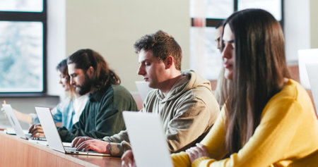 Mixed-races young adults students sitting at desks in classroom and listening to professor. Indoors. Multiethnic people studying at college. Girl with laptop. Educational concept.の写真素材