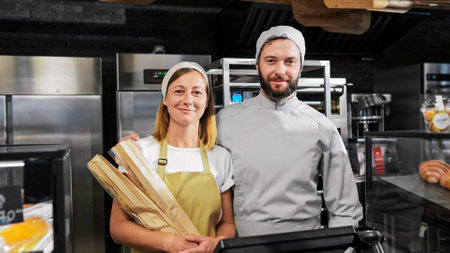 Portrait shot of Caucasian joyful couple of male and female bakers standing at bakehouse kitchen with baguettes. looking at each other and smiling to camera. Happy man and woman workers of bakeshop.の写真素材