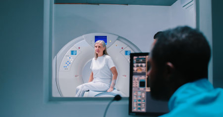 Elderly woman lies down in MRI capsule. Doctor monitors examination on computer screen. Nurse in...