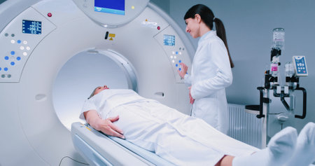 Female medical worker dressed up in gown before MRI procedure. Female doctor is gesturing and preparing patient for magnetic resonance procedure. Patient is lying at CT bed and listening to doctor.の写真素材