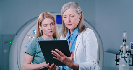 Nurse consults with head doctor. Medical workers look at results of patient examinations. Female doctor is holding tablet and showing screen to colleague. Two doctors talk at background MRI equipment.の写真素材