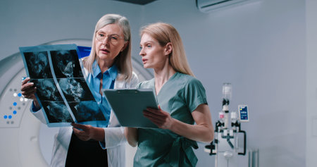 Portrait of female doctor and nurse standing at MRI room. Doctor is holding scans and talking to colleague. Nurse is holding folder and making notes with pen. Doctor and nurse analize MRI results.の写真素材