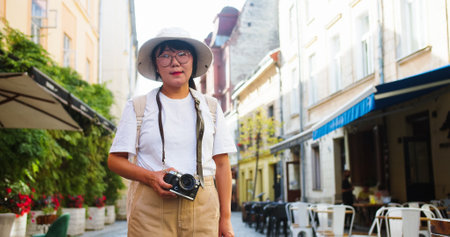 Portrait of joyful middle-aged female tourist in glasses and hat standing in town with photo camera...