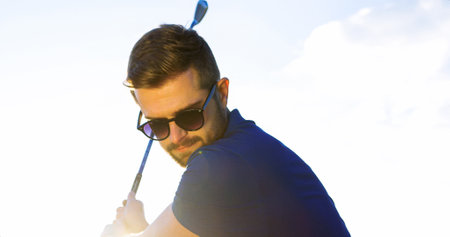 Close up of an attractive caucasian man in sunglusses making a swing with a club on the blue sky...