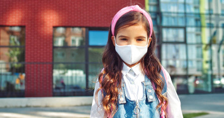 Close up portrait of nice beautiful Caucasian little girl in mask smiling in front of school. Pretty cute school kid standing with smile on face outdoor. Back to school in quarantine. Study conceptの写真素材