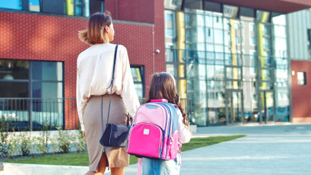 Rear of Caucasian beautiful mother and cute daughter in masks going to lessons outdoors in sun lights. Mom leads little junior student to school after quarantine. People on background. Pupil conceptの写真素材