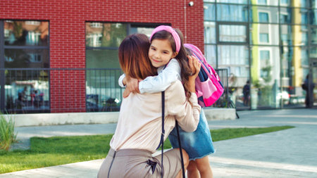 Beautiful happy Caucasian woman standing outdoor and waiting for little daughter from school. Cheerful cute girl in pink headband hugging mother after lessons and talking about classes. Family conceptの写真素材