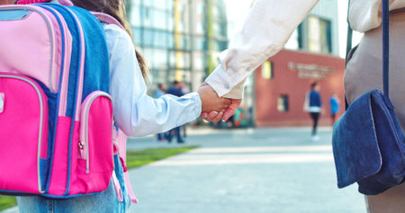 Rear of Caucasian mother holding hand of little daughter pupil with backpack and walking going to school outdoor. Close up of female parent leads girl to lessons on street. Back to school conceptの写真素材