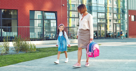 Beautiful Caucasian woman walking with little daughter pupil from school and speaking on sunny day. Joyful female walks with happy girl and talking while holding pink backpack. Conversation conceptの写真素材
