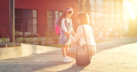 Beautiful woman speaking with cute little schoolgirl outdoors on sunny morning at schoolyard. Caucasian happy mother chatting with daughter pupil outdoors before lessons. Back to school conceptの写真素材