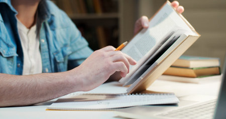 Close up of male student writing in notebook with pencil while reading textbook. Holding book and...