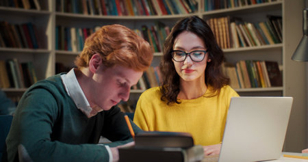 Caucasian couple of students talking and studying together in library. Male writing notes and noting important information, female explaining. Bibliotheca concept. Tutoring.の写真素材