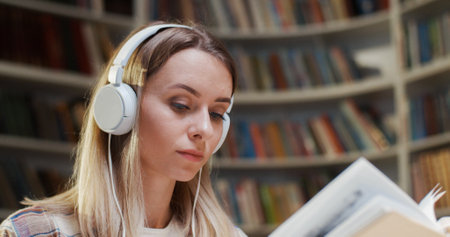 Close up of beautiful young Caucasian woman in headphones reading book and listening to music in library. Educational concept in bibliotheca. Female student listen to song and studying.の写真素材