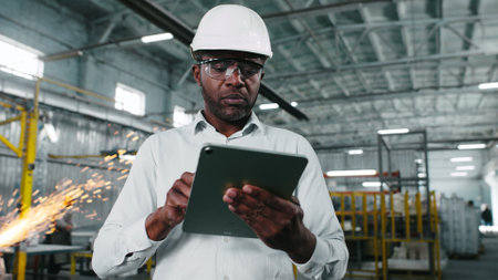 African American engineer in hard hat is looking through workshop. Man is holding gadget at construction site. Worker makes notes in tablet. Employee on background of welding sparks flying.の写真素材