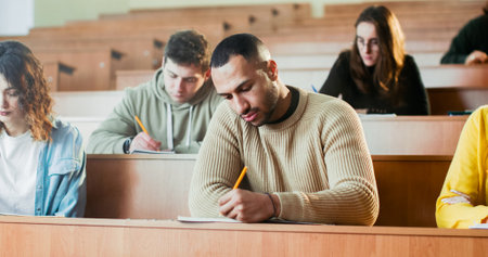 Portrait of handsome young African American guy student sitting at desk at seminar in college, noting information in copybook and smiling to camera cheerfully. Inside classroom. Guy writing down.の写真素材