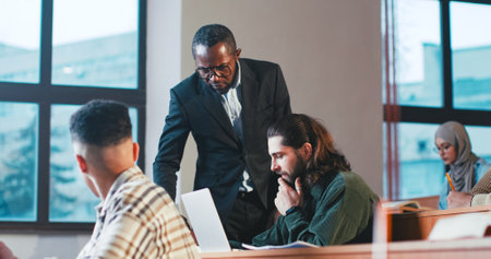 African American man professor walking the classroom during examination and helping guy student to...