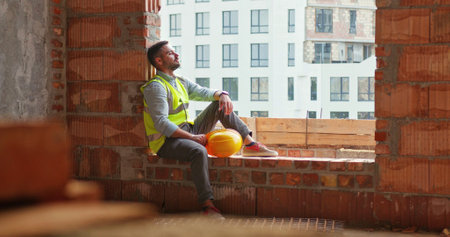 Tired Caucasian worker or foreman in vest takes off yellow helmet and wipes sweat from his forehead...