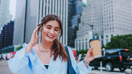 Tourist in new modern big city. Close-up of young pretty woman in headphones listening favorite music, chilling, drinking tea or coffee from glass on warm summer day, looking around cheerfully.の写真素材