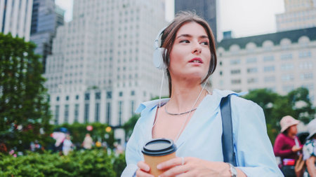 Attractive cheerful young woman enjoying music. Pretty caucasian girl walking in big beautiful city wearing headphones listening music holding cup of coffee.の写真素材
