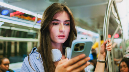 Brunette young woman listening music or podcast and chatting using wi-fi internet while traveling in underground train. Portrait of smiling lovely girl typing message on mobile phone in subway train.の写真素材