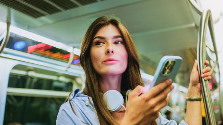 Female chatting online in mobile phone messenger watch video in metro. Charming woman wearing wireless headphones while moving in tram. Big city lifestyle, transportation.の写真素材