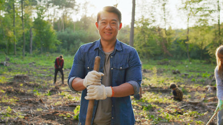Portrait of male asian eco activist wearing gloves standing with a shovel and looking at the camera while posing with multicultural people cleaning rubbish at the backgroundの写真素材