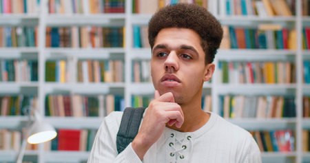 Close up of handsome Latino guy standing in library and looking at camera with thoughtful face. Portrait shot of serious male student in bibliotheca considering something. Books shelves on background.の写真素材