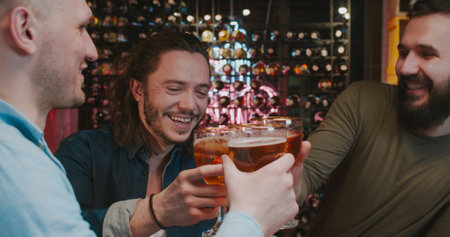 Caucasian handsome guys doing cheers gesture with beer in pub in evening and watching sport channel on TV. Friends talking and discussing football game in bar. Male cheerful fans.の写真素材